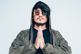 A man stands with hands pressed together in a gesture of prayer or meditation. He wears a green jacket with a hood pulled over his head and stylish round sunglasses. His expression is calm and contemplative.