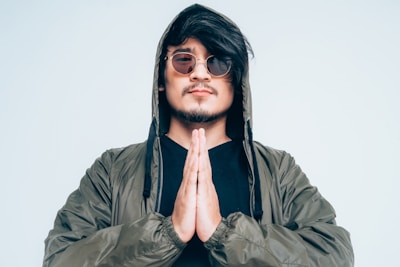 A man stands with hands pressed together in a gesture of prayer or meditation. He wears a green jacket with a hood pulled over his head and stylish round sunglasses. His expression is calm and contemplative.