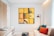 A contemporary living room with a large, vibrant artwork composed of various yellow sections featuring small details like a plant and a bicycle wheel. The space includes a white couch adorned with blue and orange pillows, next to a modern black floor lamp. A wooden coffee table displays a bowl of decorative spheres. On the left, a wooden cabinet holds a couple of books, under an inviting, soft natural light created by white curtains.