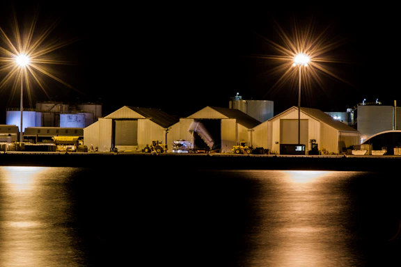 Modern industrial warehouse exterior at dusk with dramatic lighting and trucks parked nearby.