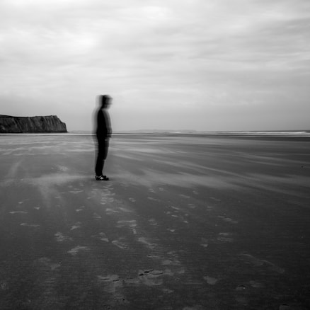 A shadowy figure appears blurred and indistinct standing on a vast, desolate beach. The overcast sky creates a moody atmosphere, with the ocean visible in the distance and a large cliff on the left side of the image. Footprints are faintly visible in the sand, adding to the eerie, isolated feel.