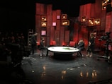 A stage setup with panel walls and warm lighting creates an intimate scene. Several people are engaged in a conversation or presentation around a large, round table. The background is decorated with red and brown wooden panels and multiple small lightboxes. A camera is set up in the corner, and an audience is visible in the darkened area.