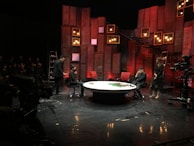 A stage setup with panel walls and warm lighting creates an intimate scene. Several people are engaged in a conversation or presentation around a large, round table. The background is decorated with red and brown wooden panels and multiple small lightboxes. A camera is set up in the corner, and an audience is visible in the darkened area.