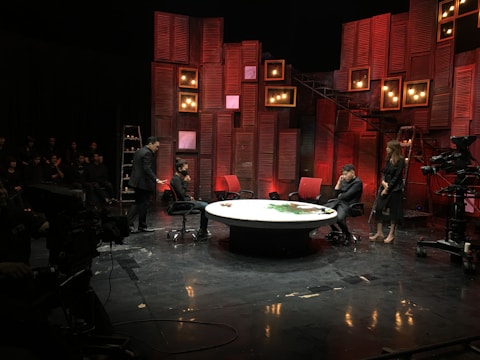 A stage setup with panel walls and warm lighting creates an intimate scene. Several people are engaged in a conversation or presentation around a large, round table. The background is decorated with red and brown wooden panels and multiple small lightboxes. A camera is set up in the corner, and an audience is visible in the darkened area.