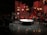A stage setup with panel walls and warm lighting creates an intimate scene. Several people are engaged in a conversation or presentation around a large, round table. The background is decorated with red and brown wooden panels and multiple small lightboxes. A camera is set up in the corner, and an audience is visible in the darkened area.