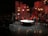 A stage setup with panel walls and warm lighting creates an intimate scene. Several people are engaged in a conversation or presentation around a large, round table. The background is decorated with red and brown wooden panels and multiple small lightboxes. A camera is set up in the corner, and an audience is visible in the darkened area.