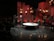 A stage setup with panel walls and warm lighting creates an intimate scene. Several people are engaged in a conversation or presentation around a large, round table. The background is decorated with red and brown wooden panels and multiple small lightboxes. A camera is set up in the corner, and an audience is visible in the darkened area.
