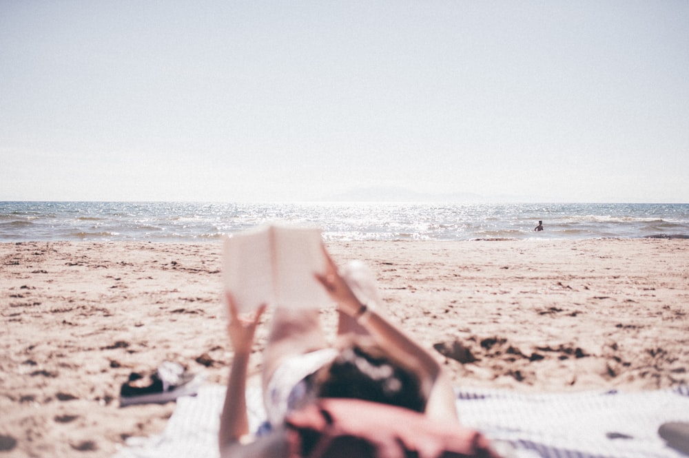 person lying on blanket while reading on beach