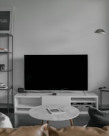 A modern living room setup featuring a large flat-screen television on a white console against a light gray wall. To the left, there is a black metal shelf with books and decorative items. In front of the TV, a round marble coffee table with a magazine on top is surrounded by a light brown leather sofa and a dark gray cushion. A minimalistic floor lamp hangs from the wall on the right.