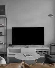 A modern living room setup featuring a large flat-screen television on a white console against a light gray wall. To the left, there is a black metal shelf with books and decorative items. In front of the TV, a round marble coffee table with a magazine on top is surrounded by a light brown leather sofa and a dark gray cushion. A minimalistic floor lamp hangs from the wall on the right.