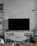 A modern living room setup featuring a large flat-screen television on a white console against a light gray wall. To the left, there is a black metal shelf with books and decorative items. In front of the TV, a round marble coffee table with a magazine on top is surrounded by a light brown leather sofa and a dark gray cushion. A minimalistic floor lamp hangs from the wall on the right.