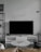 A modern living room setup featuring a large flat-screen television on a white console against a light gray wall. To the left, there is a black metal shelf with books and decorative items. In front of the TV, a round marble coffee table with a magazine on top is surrounded by a light brown leather sofa and a dark gray cushion. A minimalistic floor lamp hangs from the wall on the right.