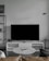 A modern living room setup featuring a large flat-screen television on a white console against a light gray wall. To the left, there is a black metal shelf with books and decorative items. In front of the TV, a round marble coffee table with a magazine on top is surrounded by a light brown leather sofa and a dark gray cushion. A minimalistic floor lamp hangs from the wall on the right.