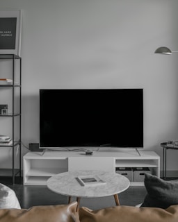 A modern living room setup featuring a large flat-screen television on a white console against a light gray wall. To the left, there is a black metal shelf with books and decorative items. In front of the TV, a round marble coffee table with a magazine on top is surrounded by a light brown leather sofa and a dark gray cushion. A minimalistic floor lamp hangs from the wall on the right.