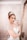 A woman with red hair is wearing a white wedding gown adorned with glittering details and a sparkling tiara. She has dramatic makeup with bold red lipstick and is posing with her hand near her face. The background features an elegant, patterned wallpaper and a large ornate mirror.