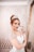 A woman with red hair is wearing a white wedding gown adorned with glittering details and a sparkling tiara. She has dramatic makeup with bold red lipstick and is posing with her hand near her face. The background features an elegant, patterned wallpaper and a large ornate mirror.