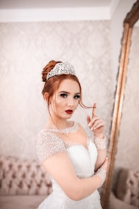 A woman with red hair is wearing a white wedding gown adorned with glittering details and a sparkling tiara. She has dramatic makeup with bold red lipstick and is posing with her hand near her face. The background features an elegant, patterned wallpaper and a large ornate mirror.