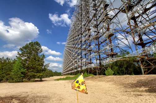 A large array of towering metal structures stands in a sandy, open area with a clear, blue sky and fluffy white clouds. To the left, there are several tall green trees. In the foreground, a rusted yellow triangular sign with a radiation warning symbol is stuck in the sand.
