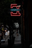 A dimly lit street scene with a prominent neon sign that reads 'ESTUPENDA' in glowing red and blue, mounted on a lamppost. The street is lined with trees and people are seen walking or standing in the background, some near a scooter. The atmosphere is urban and slightly mysterious.