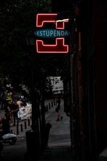 A dimly lit street scene with a prominent neon sign that reads 'ESTUPENDA' in glowing red and blue, mounted on a lamppost. The street is lined with trees and people are seen walking or standing in the background, some near a scooter. The atmosphere is urban and slightly mysterious.