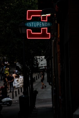 A dimly lit street scene with a prominent neon sign that reads 'ESTUPENDA' in glowing red and blue, mounted on a lamppost. The street is lined with trees and people are seen walking or standing in the background, some near a scooter. The atmosphere is urban and slightly mysterious.