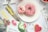 Two donuts on a white plate, one with pink icing and sprinkles, the other with white icing and red sprinkles, are placed on a distressed wooden surface. Surrounding the plate are various magazines and a notebook with a watermelon design and a motivational quote. A pink heart-shaped cookie and a bowl of strawberries are also visible.
