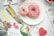 Two donuts on a white plate, one with pink icing and sprinkles, the other with white icing and red sprinkles, are placed on a distressed wooden surface. Surrounding the plate are various magazines and a notebook with a watermelon design and a motivational quote. A pink heart-shaped cookie and a bowl of strawberries are also visible.