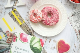 Two donuts on a white plate, one with pink icing and sprinkles, the other with white icing and red sprinkles, are placed on a distressed wooden surface. Surrounding the plate are various magazines and a notebook with a watermelon design and a motivational quote. A pink heart-shaped cookie and a bowl of strawberries are also visible.