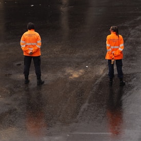 Two individuals dressed in bright orange jackets with reflective stripes stand on a wet, dark surface. The jackets have 'Protección Civil' written on the back. They appear to be observing or monitoring the surrounding area.