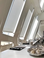 A modern interior space with large angled windows allowing ample natural light. A sleek, white countertop is set up with empty wine glasses and neatly placed napkins. A plate holds several oyster shells on ice, and there are other dining items like cutlery and glasses in the background.