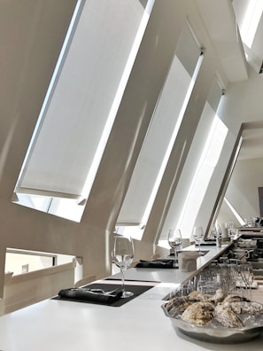 A modern interior space with large angled windows allowing ample natural light. A sleek, white countertop is set up with empty wine glasses and neatly placed napkins. A plate holds several oyster shells on ice, and there are other dining items like cutlery and glasses in the background.