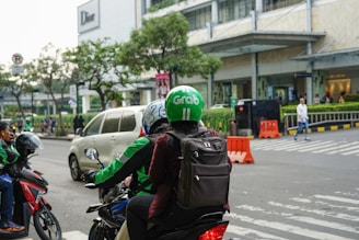 Two people wearing helmets and jackets are on a motorcycle, waiting at a pedestrian crossing near a shopping area. A white car is passing by in front of a building with a 'Dior' sign. There are orange traffic barriers and pedestrians in the background, along with roadside greenery and a city atmosphere.