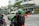 Two people wearing helmets and jackets are on a motorcycle, waiting at a pedestrian crossing near a shopping area. A white car is passing by in front of a building with a 'Dior' sign. There are orange traffic barriers and pedestrians in the background, along with roadside greenery and a city atmosphere.