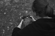 A person with tied hair is sitting near a rocky surface, holding a small camera. They are wearing a hooded sweatshirt and camo-patterned pants. Their focus appears to be on the photography task.
