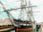 A historic sailing ship with numerous ropes and rigging, docked at a harbor with a visible wooden pier. The ship has a large black hull with elegant white detailing and an American flag. Several people are seen walking on the pier and in the background, there are a few industrial and maritime buildings.