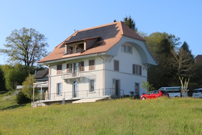 A charming two-story house with a reddish-brown tiled roof featuring solar panels is situated on a lush green lawn. The house has several windows and balconies, adding an inviting appearance. Surrounding the house are various trees and shrubs, indicating a serene, rural setting. Several cars are parked nearby, emphasizing a peaceful residential area.
