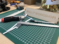 A workspace featuring a green cutting mat with measurement markings, placed on a table. Various tools such as a utility knife, caliper, and precision knife are arranged on the mat. A metal ruler lays diagonally across the surface. In the background, there's a wooden setup labeled 'WILL IT WORK(SHOP)' and a container with wrapped items.