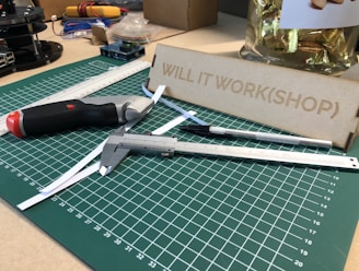 A workspace featuring a green cutting mat with measurement markings, placed on a table. Various tools such as a utility knife, caliper, and precision knife are arranged on the mat. A metal ruler lays diagonally across the surface. In the background, there's a wooden setup labeled 'WILL IT WORK(SHOP)' and a container with wrapped items.