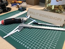 A workspace featuring a green cutting mat with measurement markings, placed on a table. Various tools such as a utility knife, caliper, and precision knife are arranged on the mat. A metal ruler lays diagonally across the surface. In the background, there's a wooden setup labeled 'WILL IT WORK(SHOP)' and a container with wrapped items.