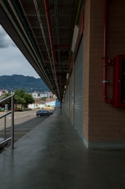 A long industrial walkway stretches alongside a building with corrugated metal doors, leading towards a distant cityscape and mountains. Overhead hang red, blue, and green pipes parallel to the walkway. A few parked vehicles are visible along with trees near the walkway's end.