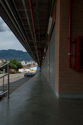A long industrial walkway stretches alongside a building with corrugated metal doors, leading towards a distant cityscape and mountains. Overhead hang red, blue, and green pipes parallel to the walkway. A few parked vehicles are visible along with trees near the walkway's end.