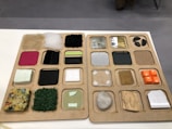 A display board showcasing a variety of different materials, each placed within individual square compartments. The materials include textiles, foam, carpet, smooth and rough stones, wood, metal, and plastic pieces. Each compartment contains a different texture and color, offering a diverse range of tactile experiences. The board is neatly organized with uniform squares arranged in rows and columns.