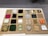 A display board showcasing a variety of different materials, each placed within individual square compartments. The materials include textiles, foam, carpet, smooth and rough stones, wood, metal, and plastic pieces. Each compartment contains a different texture and color, offering a diverse range of tactile experiences. The board is neatly organized with uniform squares arranged in rows and columns.