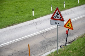 Two triangular road signs are positioned alongside a country road with a green grassy verge. The sign closer to the road depicts a pedestrian crossing symbol with '4x' written underneath. The other sign, slightly set back, shows a construction or roadwork warning symbol. The road itself shows visible cracks and wear.