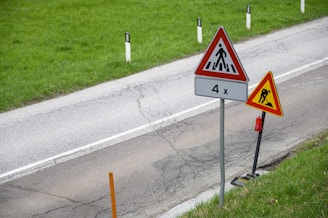 Two triangular road signs are positioned alongside a country road with a green grassy verge. The sign closer to the road depicts a pedestrian crossing symbol with '4x' written underneath. The other sign, slightly set back, shows a construction or roadwork warning symbol. The road itself shows visible cracks and wear.