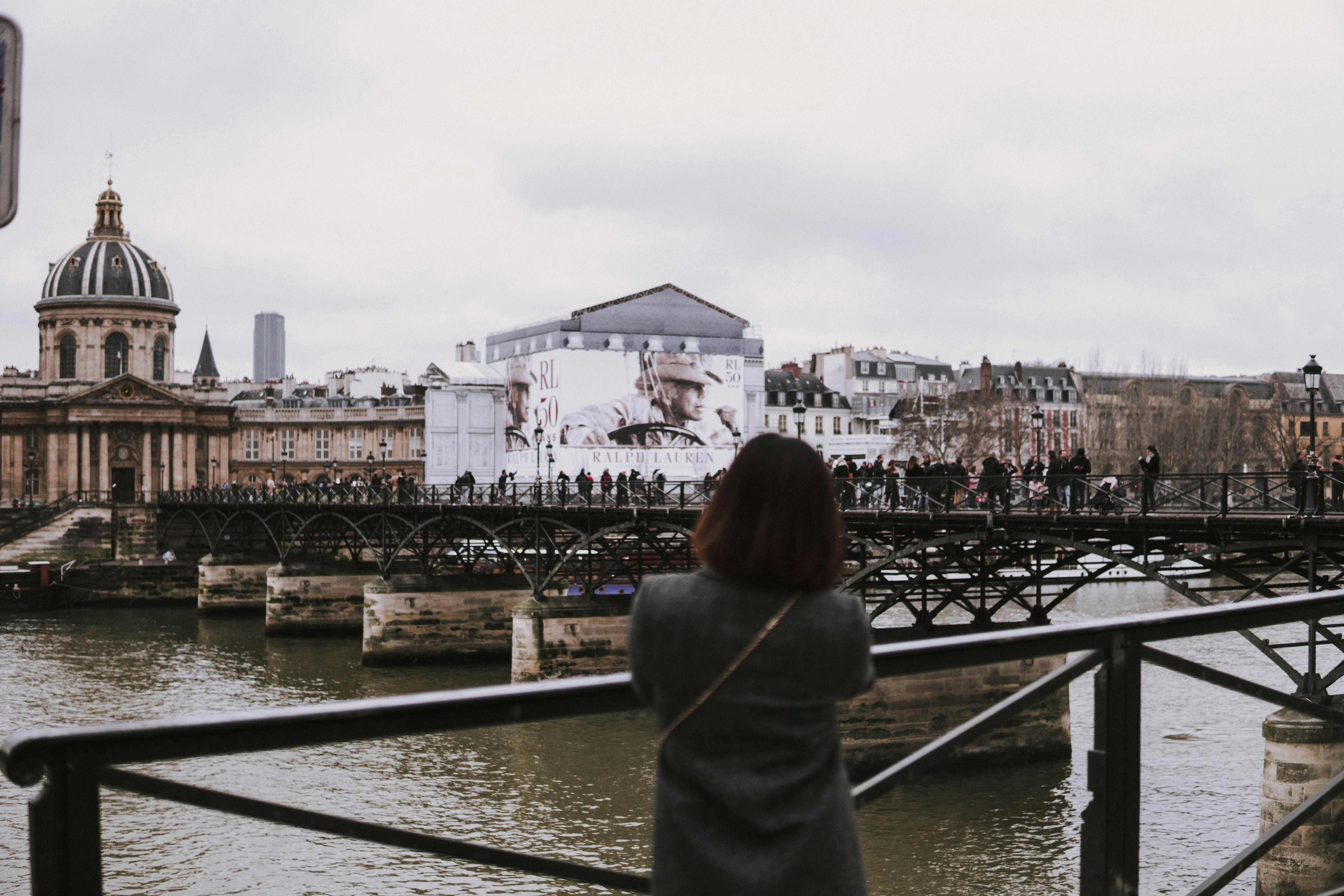 Femme en haut gris debout près des balustrades noires face au plan d’eau sous le ciel blanc pendant la journée