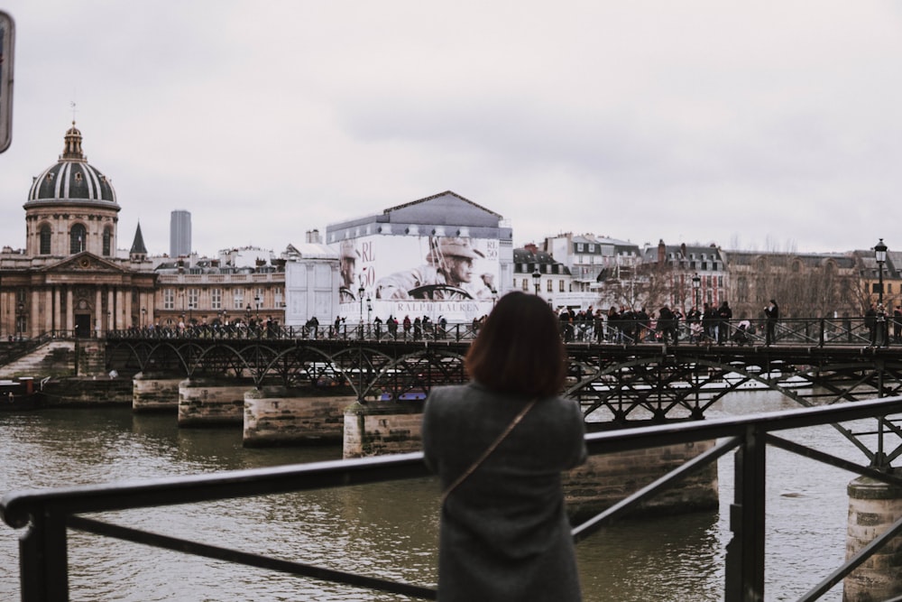 woman in gray top standing near black railings facing body of water under white sky during daytime