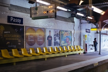 A subway station platform with a row of yellow plastic seats and advertising posters on the tiled wall. A person stands near a map or information panel, and an electronic sign displays the subway line and wait time.