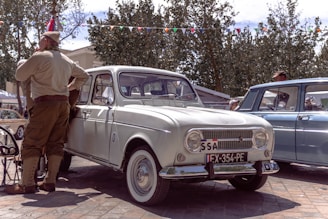 A vintage white car is parked on a street beside a man dressed in retro military-style clothing. Several other vintage cars are visible in the background, and festive bunting is strung between trees under a partly cloudy sky.