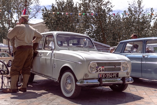 A vintage white car is parked on a street beside a man dressed in retro military-style clothing. Several other vintage cars are visible in the background, and festive bunting is strung between trees under a partly cloudy sky.